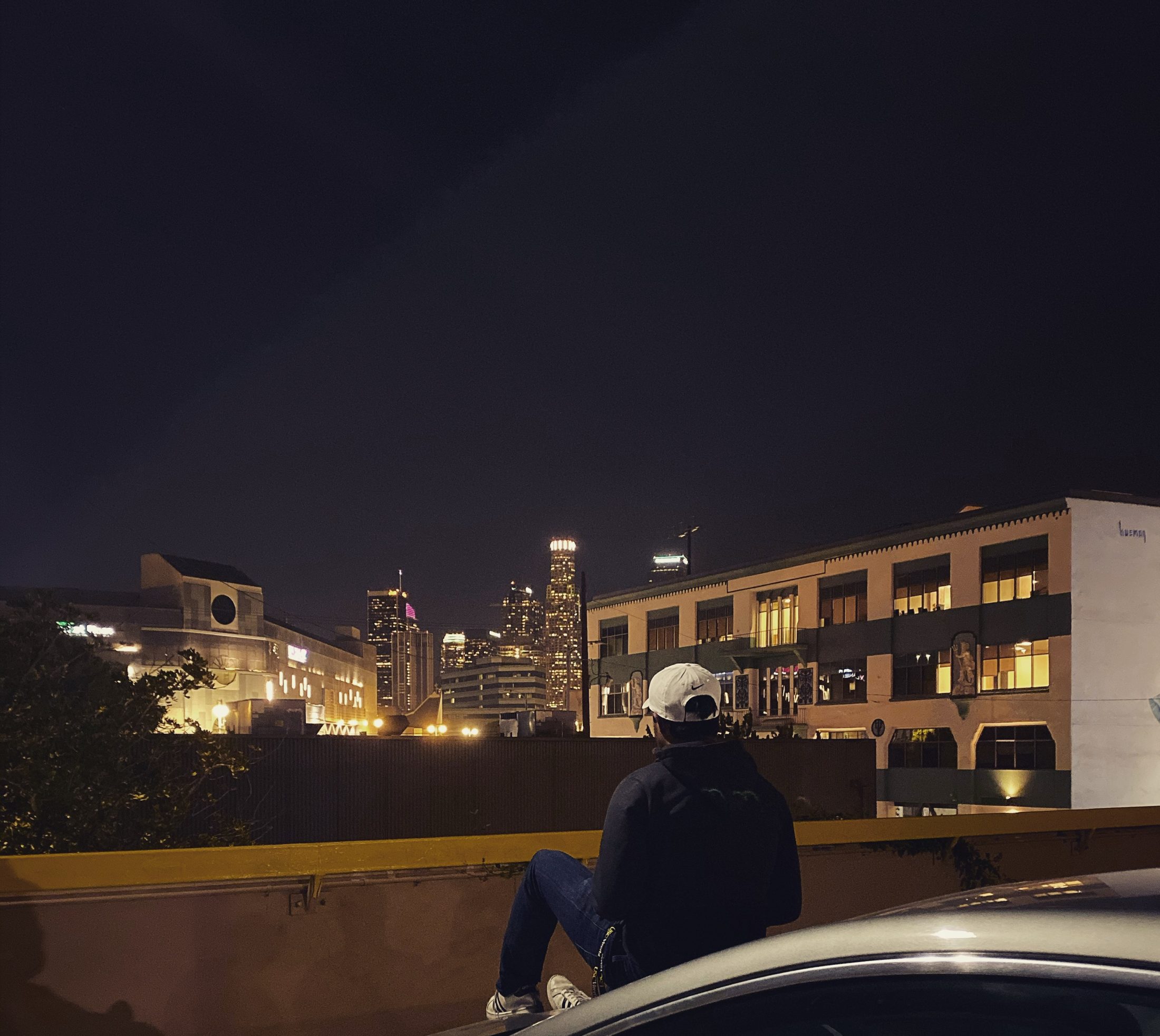Shan sitting on a car hood at night, looking out at the downtown Los Angeles skyline